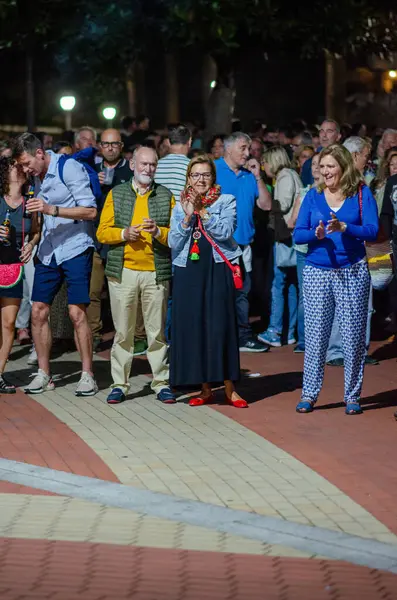 Şehir parkında düzenlenen açık hava toplantısına katılan çeşitli yetişkinlerden oluşan bir grup, halka açık bir etkinlik sırasında şenlikli bir atmosferde alkışlıyor ve iletişim kuruyor..