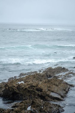 Bulutlu bir gökyüzü altında çarpışan dalgalarla vahşi kayalıklar sakin ve dramatik okyanus manzarasını yakalar. Seyahat ve doğa kavramları için idealdir..
