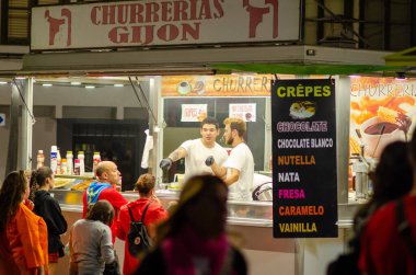 Geceleri Gijn 'deki İspanyol churros standında satıcılar canlı bir kalabalık için aperatif hazırlıyorlar. Otantik sokak yemekleri, yerel festival atmosferi.