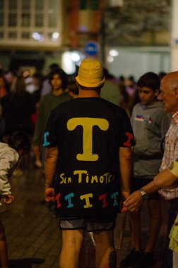 Renkli bir Aziz Timoteo tişörtü ve sarı şapkası olan bir adamın arka planı. Canlı bir gece sokak etkinliği sırasında. Etrafı şenliklerle çevrili..