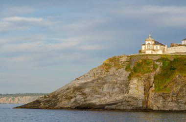 Sakin deniz sularının üzerindeki engebeli bir uçuruma tünemiş deniz feneri ve eski mimari manzarası, ılık güneş ışığıyla yıkanmış. Seyahat, doğa ve kıyı temaları için mükemmel..