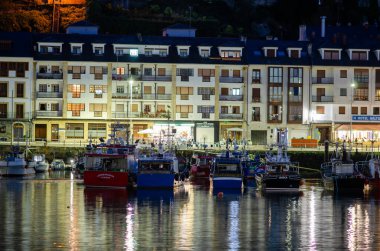 Renkli balıkçı teknelerinin durgun sularda demirlediği hareketli bir şehir limanının gece manzarası, bina ışıklarını yansıtıyor, seyahat ve turizm kavramları için mükemmel..