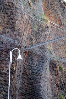 Önünde beyaz bir sokak lambası olan kayalıkların düşmesini önlemek için engebeli bir uçuruma kurulmuş metal ağların ayrıntıları. Kentsel güvenlik ve altyapı kavramı.