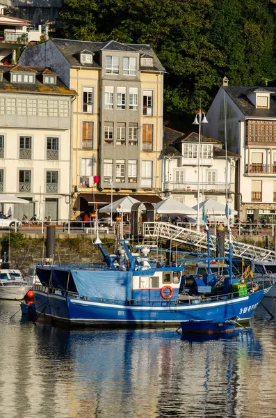 Geleneksel mavi balıkçı teknesi, canlı sahil binaları ve açık hava kafeleri ile dolu bir Avrupa sahil köyü limanına demirlemiş..