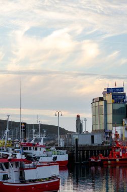 Modern balıkçı tekneleri, deniz kıyısındaki binaların yanındaki canlı bir limana demirlediler dramatik bir akşam gökyüzü ile sakin bir sahil şehri atmosferini yakaladılar..