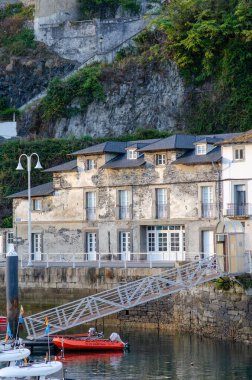 Huzurlu bir marinada soyulmuş yüzüyle otantik tarihi liman binası rıhtıma demirlemiş tekneler ve arka planda yemyeşil kayalık yamaçlar..