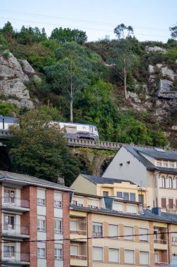 Modern tren, kayalık bir yamaçtan geçerek, kentsel konutlara ve yoğun yeşilliğe tepeden bakan, keşif ve şehir yaşamının zıtlığını çağrıştıran bir viyadük boyunca seyahat eder..