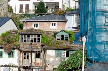 Terk edilmiş, çatıda yeşil bitkiler ve kırık camlar büyüyen bir ev, modern binalarla ve kentsel çürümeyi gösteren bir restorasyon alanıyla çelişiyor..