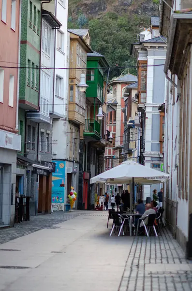Çevresi canlı tarihi mimari ve yerel dükkanlarla çevrili, sakin bir şehir atmosferini çağrıştıran açık bir kafede oturan insanların olduğu eski bir cadde..