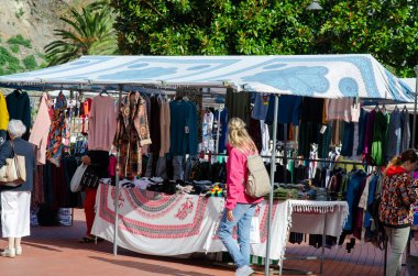 Kanopi altındaki giyim mağazalarına göz atan insanların olduğu hareketli bir açık hava piyasa sahnesi. Güneşli bir günde günlük alışverişi, pazar atmosferini ve şehir hayatını ön plana çıkarıyor.