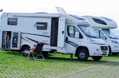 Otlak bir kamp yerine kurulmuş rahat bir karavan. Açık sandalyesi var. Rahatlatıcı bir tatil hayat tarzı ve karavan özgürlüğü sergiliyor..