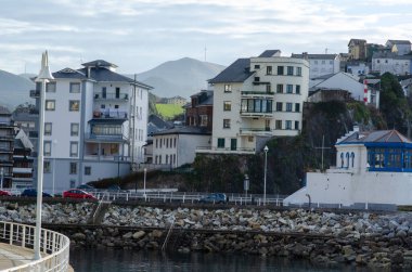 Resimli sahil kasabası, kayalık bir rıhtıma yakın modern konut binaları, arka planda dingin dağlar ve gezinti güvertesindeki arabalar, seyahat veya mimari kavramlar için ideal..