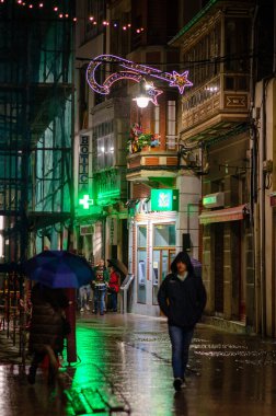 Yayalar, canlı bir Avrupa şehrinin caddesinde, neşeli Noel ışıklarının altında yürürler, ıslak kaldırımları, kentsel tatil atmosferini yansıtırlar..