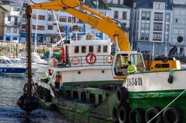 Bir tarama gemisi yoğun bir marinada çalışır, liman derinliğini ağır makinelerle korur. Liman işçileri ve ekipmanlar endüstriyel deniz faaliyetlerini vurguluyor.
