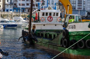 Vinçli ve mürettebatlı bir tarama gemisi, arka planda yatları ve belediye binaları olan yoğun bir marinada çalışıyor. Deniz sanayisi ve liman operasyonlarını resmetmek için ideal.