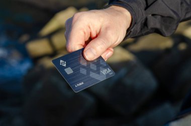 Adam elinde bir Binance Visa kredi kartı tutuyor. Şifreli para evlat edinme ve dijital finansı sembolize ediyor. Açık hava sahnesi teknolojik yenilik ve finansal özgürlüğü vurguluyor.