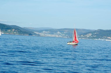 Canlı kırmızı bir yelkenli, kıyı kenti yakınlarındaki sakin mavi sularda süzülüyor. Çerçevelenmiş tepeler ve modern asma köprü, özgürlük ve macera çağrıştırıyor..