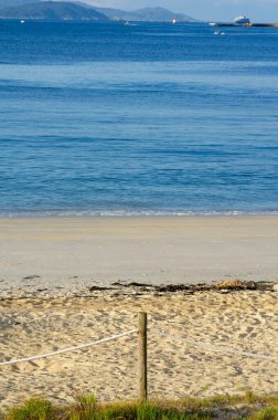 Sakin bir sahil manzarası. Kumlu bir sahil. Halat çitleri, yumuşak deniz dalgaları ve açık mavi gökyüzü altında uzak dağlar. Tatil ve rahatlama temaları için mükemmel.
