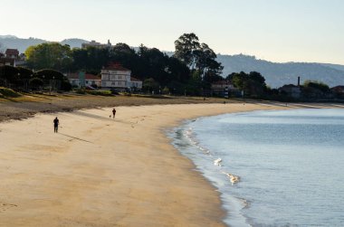 Sabahın erken saatlerinde sakin kumlu bir sahil manzarası. Hafif dalgalar ve sahil boyunca yürüyen birkaç insan. Rahatlamak ve seyahat etmek için mükemmel bir yer..