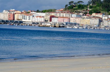 Canlı bir sahil kasabasının panoramik manzarası marinada demirli yatlar, sakin mavi sular ve bulutlu bir gökyüzünün altındaki yamaçtaki konut binaları..