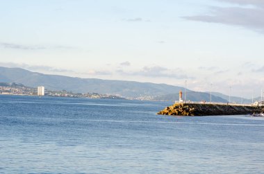 Kızıl feneri, sakin denizi, uzak şehir binaları ve yeşil dağları olan bir sahil iskelesinin sakin manzarası seyahat veya manzara kavramları için mükemmeldir..