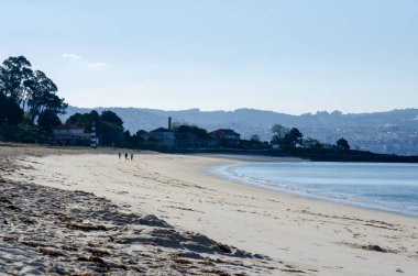 Yumuşak kumları ve yumuşak dalgaları olan sakin bir sahil, birkaç insan geziniyor, sakin bir sabah atmosferi, uzak bir şehir. Seyahat ve rahatlama temaları için mükemmel.