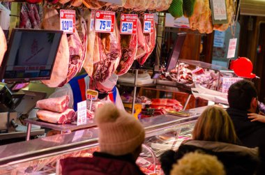 Canlı İspanyol jamonu sergilenen yerel şarküteri müşterilere hizmet eden özenli kasap ve fiyat tabelaları. Otantik pazar yeri yemek kültürü sahnesi.