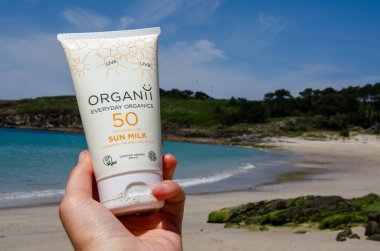 Hand holding SPF 50 sunscreen lotion against a scenic beach backdrop, evoking summer vacation, skincare routines, and UV protection during outdoor activities.