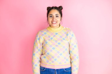 İzole edilmiş pembe arka plan üzerine sıradan bir süveter giyen genç ve güzel bir kadın. Mutlu bir yüzü olan ve kendine güvenen bir gülümsemeyle dişlerini gösteren.