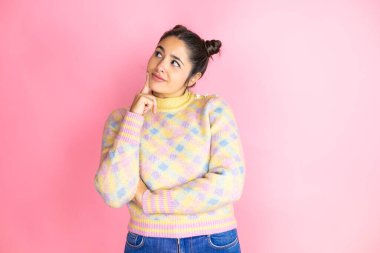 İzole edilmiş pembe arka plan üzerine sıradan süveter giyen genç ve güzel bir kadın. Mutlu bir yüz. Elle çeneyi sorgulayan, kafa karıştırıcı bir fikir.