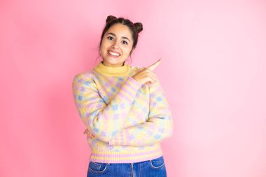 İzole edilmiş pembe arka planda rahat bir süveter giyen genç ve güzel bir kadın. Gülümsüyor ve parmaklarını kenarı işaret ediyor.
