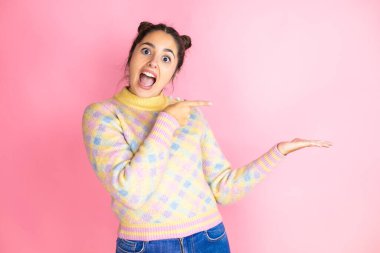 Genç, güzel bir kadın pembe arka plan üzerine günlük süveter giyiyor. Şaşırdı ve elindeki bir şeyi gösterip işaret ediyor.