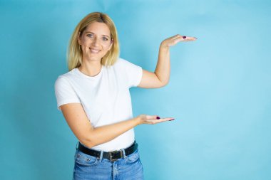 İzole edilmiş mavi arka plan jestlerinin üzerine gündelik tişört giyen genç ve güzel bir kadın. Ellerinde büyük ve büyük beden işareti, ölçü sembolü var. Gülümsüyor. Ölçüm konsepti.