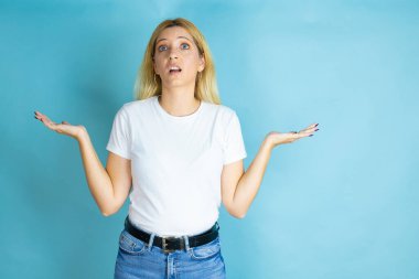 İzole edilmiş mavi arka plan üzerine günlük tişört giyen genç ve güzel bir kadın. Kolları ve elleri havada, kafası karışmış bir ifade.