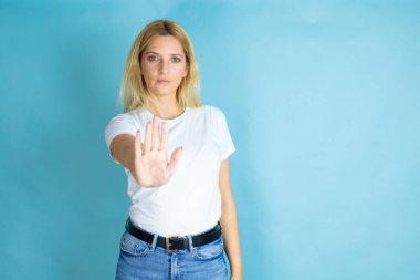 Genç ve güzel bir kadın, izole edilmiş mavi arka plan üzerine gündelik tişört giyiyor ve avuç içiyle şarkı söylemeyi kesiyor..