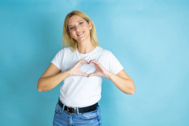 Genç, güzel bir kadın, izole edilmiş mavi arka planda günlük tişört giyiyor. Aşkla gülümsüyor. Elleriyle kalp sembolü ve şekli gösteriyor.