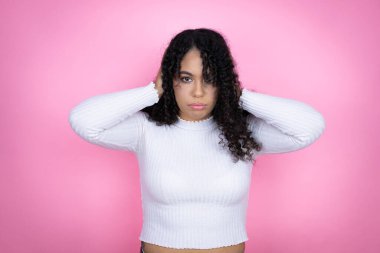 Pembe arka planda günlük süveter giyen Afro-Amerikalı bir kadın. Elleri başının üstünde yorgun ve sıkılmış görünüyor.