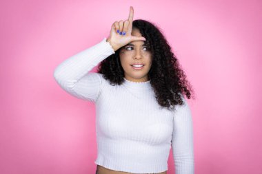 Afro-Amerikalı kadın pembe arka plan üzerine günlük süveter giyiyor. Alnında parmakları olan insanlarla dalga geçiyor. Ezik hareketler yapıyor, alay ediyor ve hakaret ediyor..