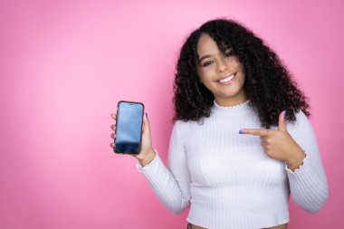 Afro-Amerikalı kadın pembe arka plan üzerine günlük süveter giyip elinde akıllı telefon ekranı tutarak mutlu bir şekilde eli ve parmağı işaret ediyor.