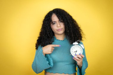 African american woman wearing casual sweater over yellow background serious holding and pointing a clock clipart