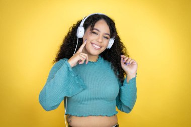Sarı arka plan üzerine günlük süveter giyen Afrikalı Amerikalı kadın kulaklık takıyor, gülümsüyor ve parmaklarıyla ve ağzıyla işaret ediyor.