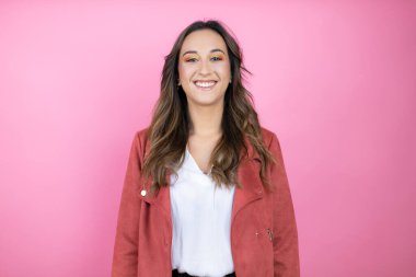 İzole edilmiş pembe arka planda rahat bir ceket giyen genç ve güzel bir kadın. Mutlu bir yüzü var. Kendine güvenen bir gülümsemeyle dişlerini gösteriyor.