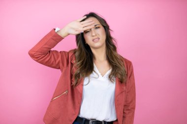 İzole edilmiş pembe arka planda rahat ceket giyen genç ve güzel bir kadın. Hastalık ve ateş için alnına dokunuyor. Grip ve soğuk algınlığı için.