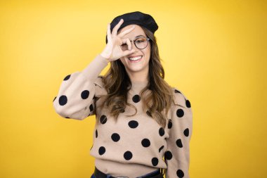 Genç, güzel esmer kadın Fransız beresi takıyor ve sarı arka planda gözlük takıyor. Gülümseyen yüzüyle, gözleriyle parmaklarından bakıyor.