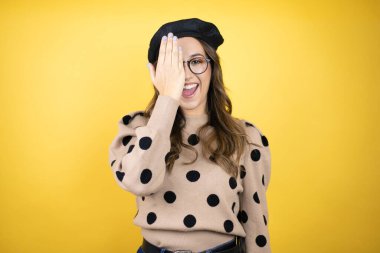 Genç, güzel esmer kadın Fransız beresi takıyor ve sarı arka planda gözlük takıyor. Bir gözünü eliyle kapatıyor. Yüzünde kendine güvenen bir gülümseme ve sürpriz bir duygu var..