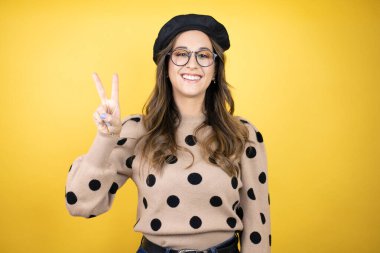 Genç, güzel esmer kadın Fransız beresi takıyor ve sarı arka planda gözlük takıyor. İki numarayı işaret ediyor ve gülümsüyor. Kendinden emin ve mutlu.
