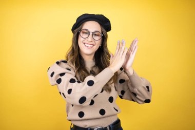 Genç, güzel esmer kadın, Fransız beresi takıyor ve sarı arka planda gözlük takıyor. Alkışlıyor, alkışlıyor, mutlu ve neşeli, gururlu elleri birleştiriyor.