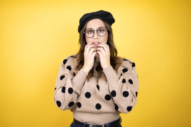 Fransız beresi takan, sarı arka planda gözlük takan genç ve güzel bir esmer kadın gergin görünüyor ve elleri ağızdan tırnak yiyor. Endişe sorunu.