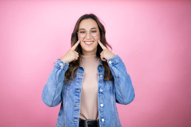 Soyutlanmış pembe arka planda kot ceket giyen genç ve güzel bir kadın mutlu ve heyecanlı. Kollarını kaldırmış, gülümseyen ve başarı için çığlık atan kazanan bir jest yapıyor..