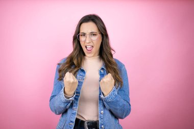 Soyutlanmış pembe arka planda kot ceket giyen genç ve güzel bir kadın mutlu ve heyecanlı. Kollarını kaldırmış, gülümseyen ve başarı için çığlık atan kazanan bir jest yapıyor..